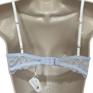 May include: A pale blue bra with white straps and a delicate lace design. The bra has adjustable straps and a hook-and-eye closure. A tag with product information hangs from the bra.