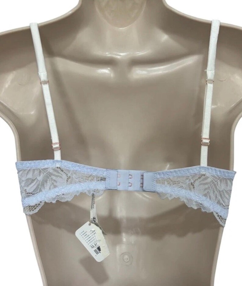 May include: A pale blue bra with white straps and a delicate lace design. The bra has adjustable straps and a hook-and-eye closure. A tag with product information hangs from the bra.