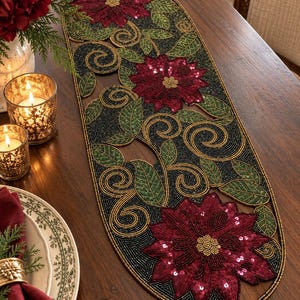 May include: An oval table runner featuring intricate beadwork in a floral pattern. The design includes deep red poinsettia flowers with sequin accents, surrounded by green beaded leaves and gold swirling vines set against a black beaded background.