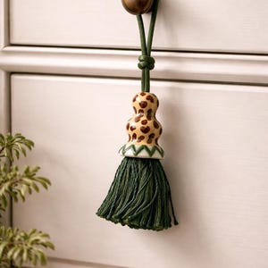 Puede incluir: Un tirador decorativo con un pomo de latón y un cordón verde. El tirador presenta una cuenta de cerámica con un diseño de estampado de leopardo y una borla verde oscuro. El tirador está sujeto a un cajón blanco.
