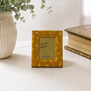 May include: A rectangular mustard yellow picture frame with a glossy finish and a repeating pattern of light yellow leaves. The center insert features the text Fashion Frames, The Bucklers, Inc., Fifth Avenue, New York in black script lettering.
