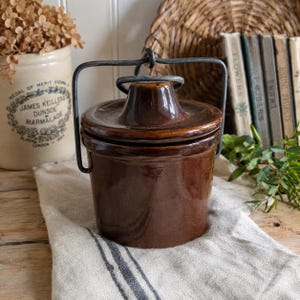 May include: A dark brown glazed ceramic crock with a matching lid and a black metal swing handle. The cylindrical container features a smooth, glossy finish and a central knob on the lid, providing a functional design for storage and kitchen use.