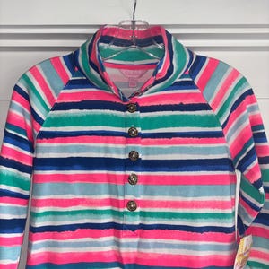 May include: A long-sleeved shirt with a colorful striped pattern in shades of pink, blue, green, and white. The shirt features a collared neckline and is fastened with decorative gold buttons. The garment is hanging on a clear hanger.