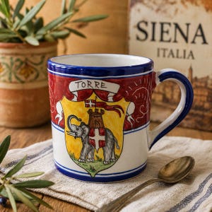 May include: A ceramic mug with a blue handle and rim, featuring a colorful crest design. The crest includes an elephant, a tower, and the word "TORRE" on a banner. The background includes the text "SIENA ITALIA".