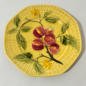 May include: Yellow ceramic plate with a woven texture and a raised design of fruit and leaves. The fruit is a deep red with yellow highlights, and the leaves are green. Two yellow flower accents are also present.