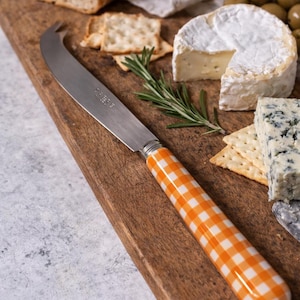 May include: A wooden cutting board with a cheese knife featuring an orange and white checkered handle. The board holds brie cheese, blue cheese, crackers, olives, and a sprig of rosemary. A loaf of bread is also present.