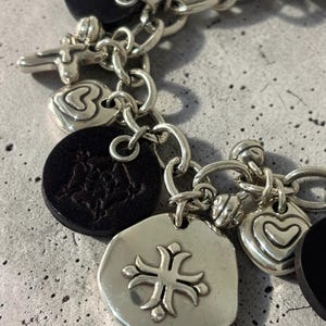 May include: Silver charm bracelet featuring a variety of pendants. Charms include a cross, hearts, and a circular pendant with a floral design. Black leather-like circular charms are also present. The bracelet is on a grey surface.