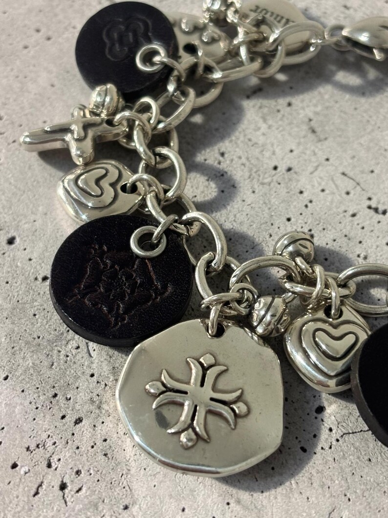 May include: Silver charm bracelet featuring a variety of pendants. Charms include a cross, hearts, and a circular pendant with a floral design. Black leather-like circular charms are also present. The bracelet is on a grey surface.