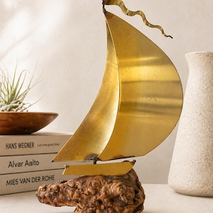 Puede incluir: Una escultura de velero de latón con una gran vela y una bandera ondulada. El barco está montado sobre una base de madera texturizada. Este artículo decorativo es adecuado para una estantería o mesa.