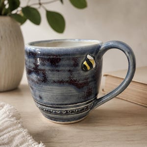 Peut inclure: Une tasse en céramique faite à la main avec un vernis brillant tacheté bleu et brun foncé. La tasse présente une petite décoration en relief en forme de bourdon jaune, noir et blanc sur le côté, avec une rangée de points circulaires en relief.