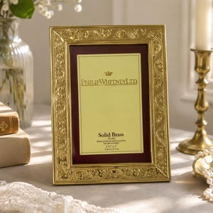 May include: A rectangular solid brass picture frame with an ornate embossed floral border. The center insert features the text PhilipWhitneyLtd, Solid Brass, and the dimensions 3.5 inches by 5 inches printed on a light yellow background.