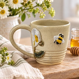 May include: A beige ceramic mug with a textured, ribbed base and a large handle. The mug features a raised, three-dimensional sculpted white calla lily with a yellow center and a green stem, alongside a small, detailed bumblebee with yellow and black stripes.