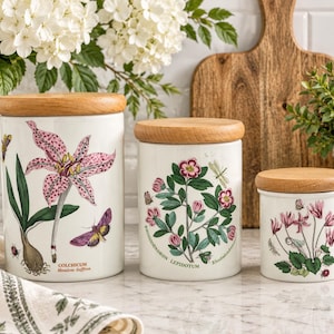 May include: A set of three white ceramic canisters with light wood lids, each featuring botanical illustrations. The largest shows a pink Colchicum, the middle a Rhododendron Lepidotum, and the smallest a Cyclamen, all with insects.