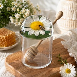 May include: A clear glass honey jar with a wooden honey dipper, resting on a wooden cutting board. The jar's lid features a large daisy with a bee. A honeycomb and flowers are in the background.