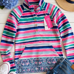 May include: A colorful striped pullover with a high collar and a kangaroo pocket. The shirt features horizontal stripes in pink, blue, green, and white. A pair of white sneakers and blue jeans are next to the shirt. A straw hat with a navy blue band is also in the image.
