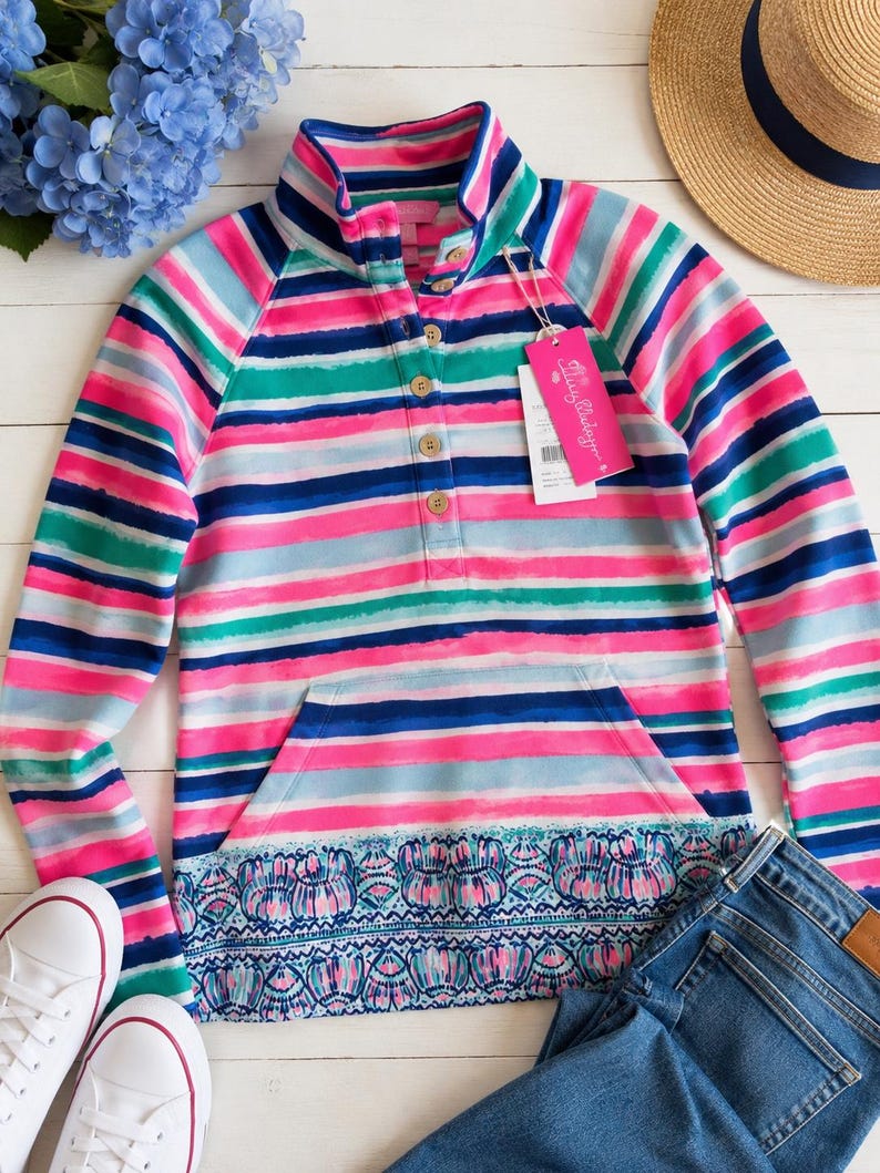 May include: A colorful striped pullover with a high collar and a kangaroo pocket. The shirt features horizontal stripes in pink, blue, green, and white. A pair of white sneakers and blue jeans are next to the shirt. A straw hat with a navy blue band is also in the image.