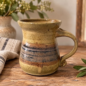 May include: A ceramic mug with a flared rim and a curved handle, featuring a speckled glaze in earthy tones of olive green, tan, and dark blue. The body of the mug has a rounded shape with horizontal bands of color and a smooth, glossy finish.