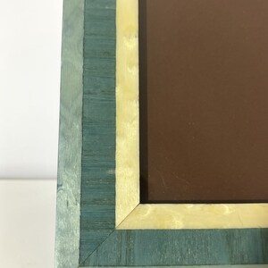 May include: A rectangular picture frame with a teal and cream-colored wood grain design. The frame has a brown interior and is set against a white background. The frame's corner is visible, showcasing the layered wood construction.