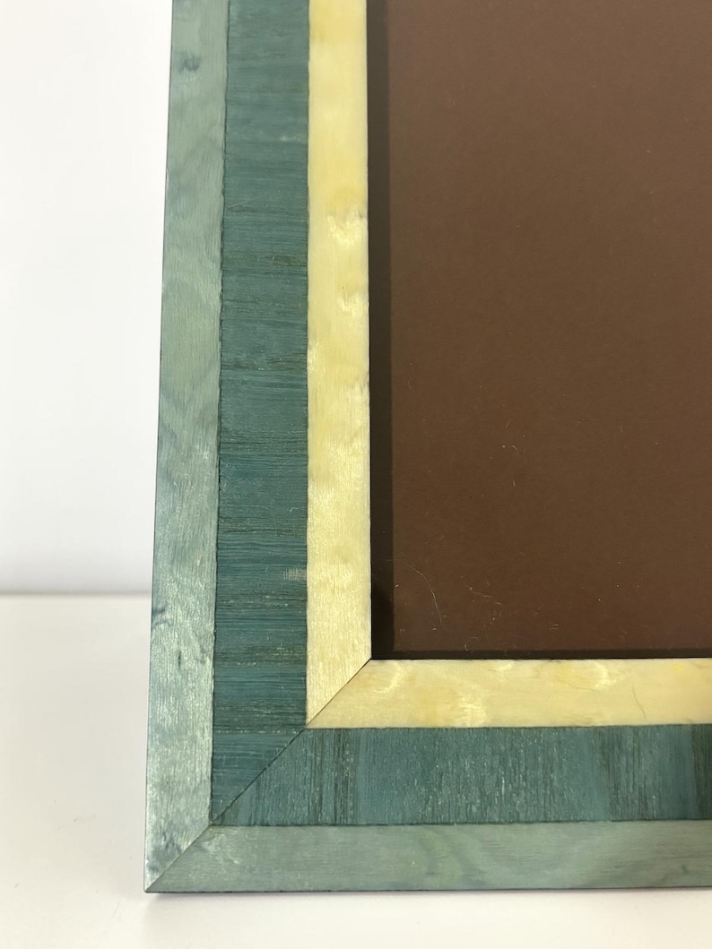May include: A rectangular picture frame with a teal and cream-colored wood grain design. The frame has a brown interior and is set against a white background. The frame's corner is visible, showcasing the layered wood construction.