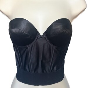 May include: A black strapless corset top with a sweetheart neckline. The bodice is made of a shiny, satin-like material with vertical seams for structure. A wide, ribbed band sits at the waist. The top is displayed on a mannequin.