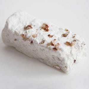 May include: A rectangular block of white nougat candy with a textured, uneven surface. The confection is topped with scattered, small, light brown pieces of chopped nuts embedded into the top layer of the soft, white, aerated sugar base.