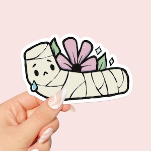 May include: A sticker featuring a cartoon illustration of a bandaged foot with a sad face and a single tear. A pink flower and green leaves are nestled in the bandages. The sticker has a white border and is held by a hand.