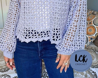 Puede incluir: Una blusa de crochet de color lila claro con un corte holgado y mangas largas. La blusa tiene un delicado patrón de calado y un dobladillo festoneado.