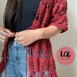 May include: A red crochet cardigan with a lacy pattern and a tie closure. The cardigan is worn over a black tank top and blue jeans. The text "LCK CROCHET" is visible in a pink circle.
