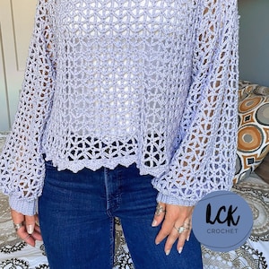 Puede incluir: Una blusa de crochet de color lila claro con un corte holgado y mangas largas. La blusa tiene un delicado patrón de calado y un dobladillo festoneado.