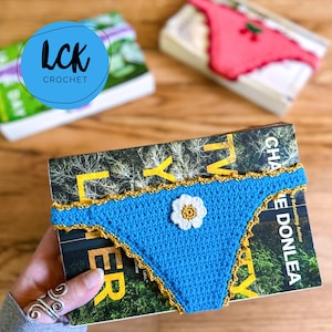 May include: A blue crochet book cover shaped like underwear, with a white daisy accent. The cover has a gold trim and is wrapped around a book. The image also shows two other books with crochet covers.