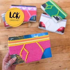 May include: A pink crochet book cover shaped like underwear with yellow and white accents. The cover is held by a hand with a silver ring. Other book covers with underwear designs are in the background. The logo "LCK CROCHET" is in the image.