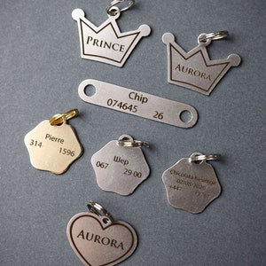 May include: A collection of silver and gold dog tags with engraved text. The tags feature different shapes, including crowns, stars, hearts, and rectangles. The text includes names, numbers, and dates.
