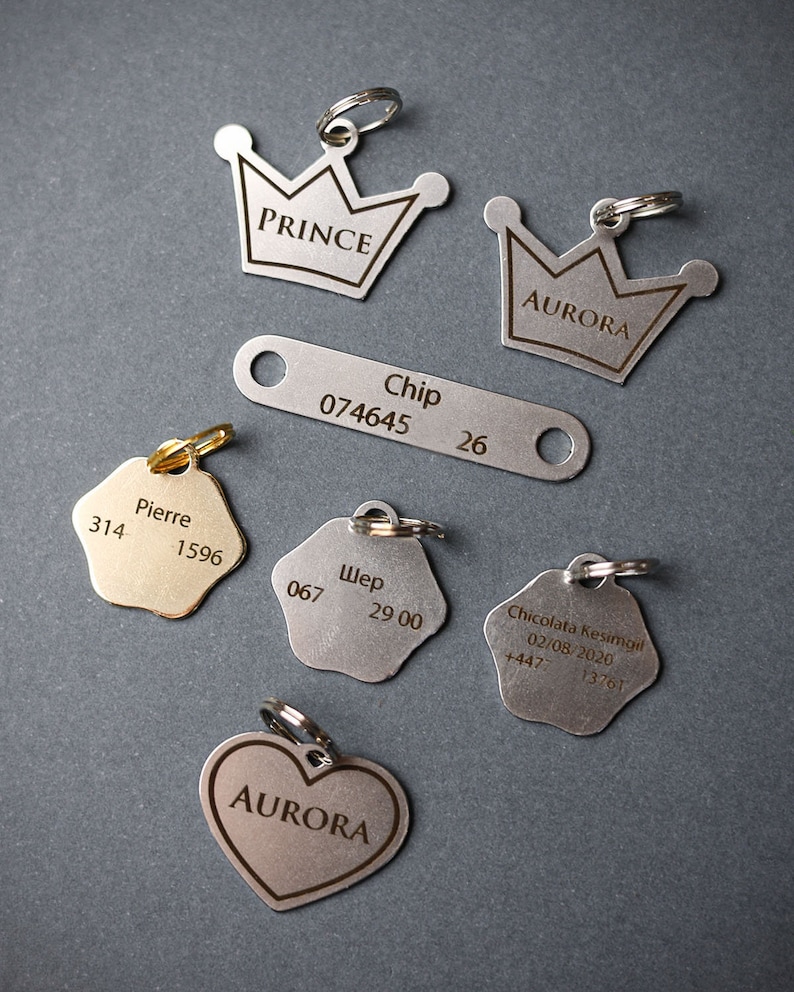 May include: A collection of silver and gold dog tags with engraved text. The tags feature different shapes, including crowns, stars, hearts, and rectangles. The text includes names, numbers, and dates.