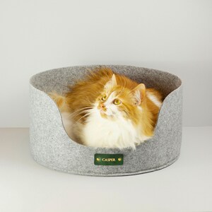 Puede incluir: Una cama para gatos de fieltro gris con un gato marrón y blanco dentro. La cama tiene una etiqueta de cuero verde con el nombre "Casper" en ella.
