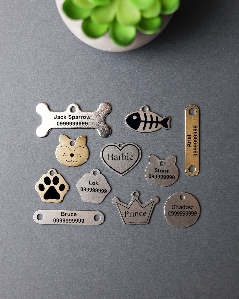 May include: Assortment of silver and gold pet ID tags with various shapes and designs, including a bone, fish, paw print, heart, crown, and cat. Each tag has a name and phone number engraved on it.  The names include Jack Sparrow, Bruce, Loki, Barbie, Storm, Ariel, Prince, and Shadow.