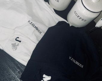 Puede incluir: Dos camisetas blancas con bordados de números romanos negros. Las camisetas tienen un parche blanco con una letra en la manga. Las camisetas están dispuestas sobre una superficie de madera oscura. El texto en las camisetas dice "V.XXILMMXIX".