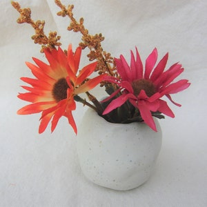 May include: A white ceramic vase with a textured surface holds two artificial flowers. One flower is orange with a brown center and the other is pink with a brown center.