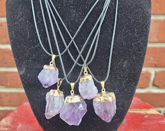 Könnte beinhalten: Sechs Amethyst-Kristall-Anhänger an schwarzen Kordeln. Jeder Anhänger ist ein roher, unregelmäßig geformter Amethyst-Kristall mit einer goldfarbenen Kappe. Die Anhänger werden an einem schwarzen Mannequin ausgestellt.