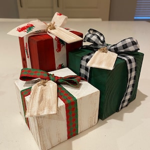 May include: Three wooden gift boxes in red, green, and white, each adorned with a decorative ribbon and a blank tag. The red box has a ribbon with a Christmas tree and truck pattern. The green box has a black and white checkered ribbon.