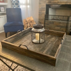 May include: A rustic wooden tray with black metal handles, featuring a glass candle lantern with a wire cage. The tray is sitting on a wooden table with a rug underneath.