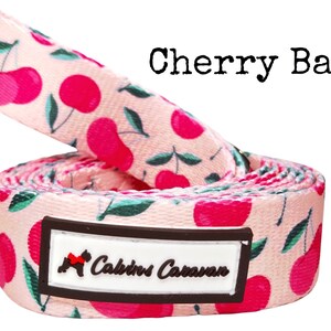 May include: Pink dog leash with a cherry print and the text "Calico's Caravan" on a brown label.