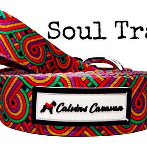 May include: A colorful dog collar with a geometric pattern in shades of red, yellow, green, purple, and black. The collar has a silver buckle and a white tag with the text "Calvin's Caravan" in black.