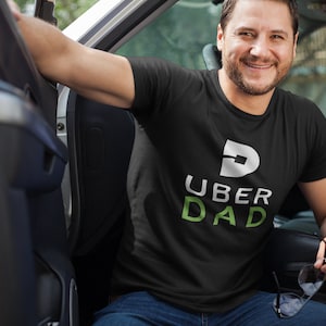 May include: Black t-shirt with the text "UBER DAD" in white and green. The shirt features a stylized "D" logo above the text. A man is sitting in a car, wearing the shirt and smiling.