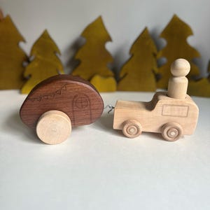May include: A wooden toy truck pulling a wooden camper trailer. The truck has a small wooden figure in the driver's seat. The camper has a small window and a door. The toys are painted in natural wood tones.
