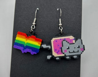 May include: A pair of dangle earrings featuring a rainbow heart and a pixelated cat with a slice of toast. The cat is wearing a pink frosting and sprinkles.