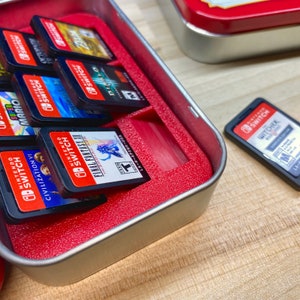 May include: A silver tin with a red interior holds several Nintendo Switch game cartridges. The cartridges are black with red and white labels, featuring game titles like "Final Fantasy XII" and "Diablo." A single cartridge, "The Witcher," rests outside the tin.