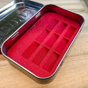 May include: An open, rectangular metal tin with a red interior. The interior features a textured, red insert with multiple rectangular compartments. The tin's lid is open, revealing the interior. The tin is on a wooden surface.