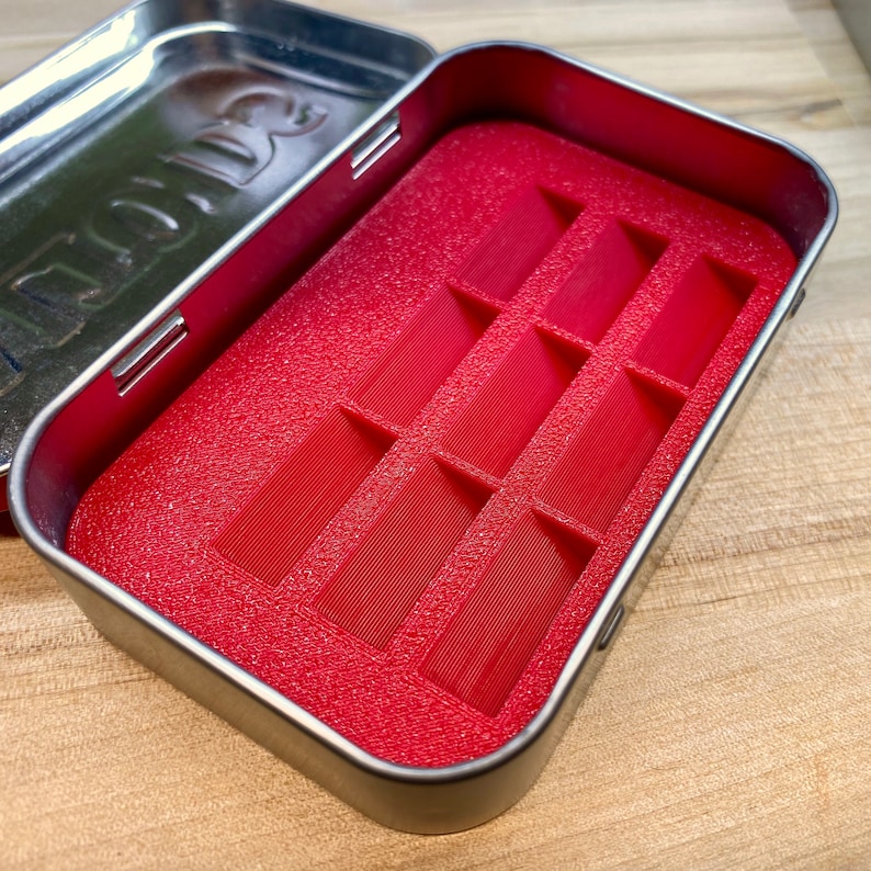 May include: An open, rectangular metal tin with a red interior. The interior features a textured, red insert with multiple rectangular compartments. The tin's lid is open, revealing the interior. The tin is on a wooden surface.