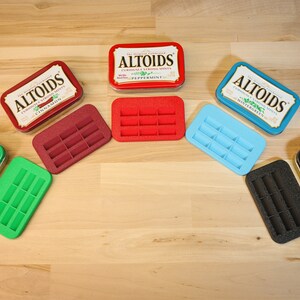 May include: An assortment of Altoids mint tins and colorful, rectangular trays. The tins are red, green, blue, and silver, with text labels. The trays are red, green, blue, burgundy, and black, each with ten rectangular slots.