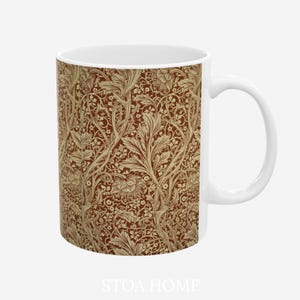 May include: A white ceramic mug featuring a repeating pattern of beige, stylized floral and leafy botanical motifs set against a deep terracotta brown background. The mug has a smooth, cylindrical body with a curved handle on the right side.